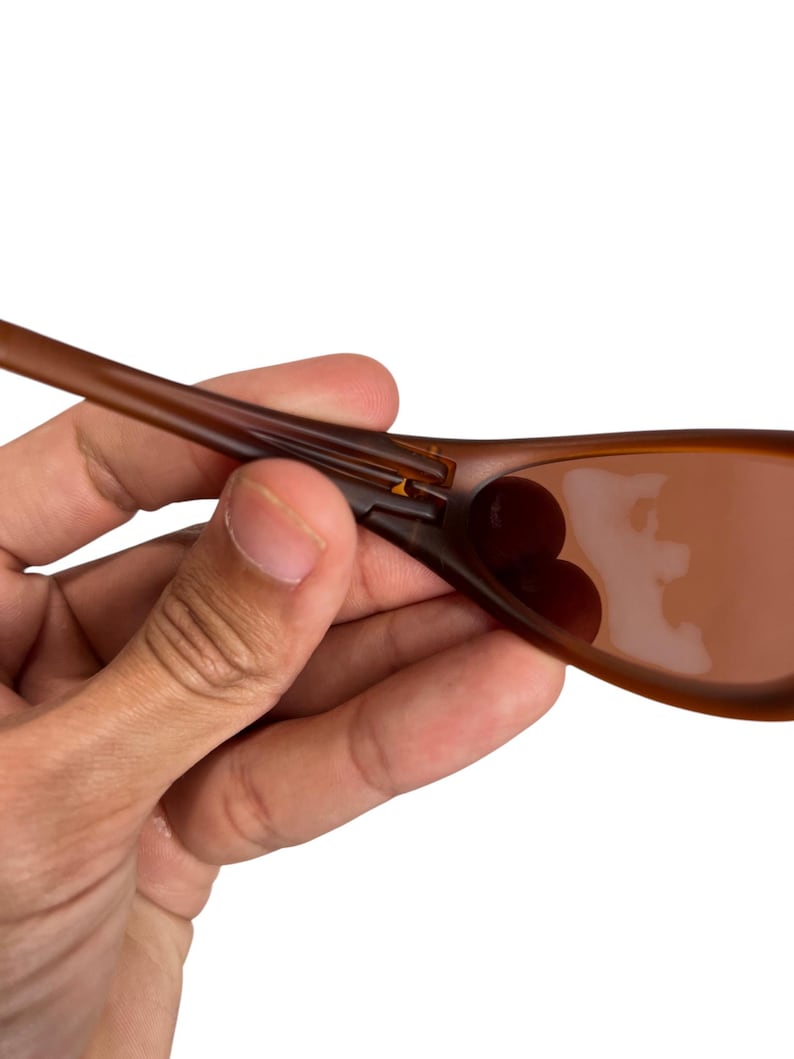 May include: Brown sunglasses with a curved frame and tinted lenses. The sunglasses are held by a hand, showcasing the frame's design and lens color. The lenses appear to be a darker shade of brown.