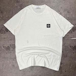 Puede incluir: Una camiseta blanca de manga corta y cuello redondo con un pequeño parche cuadrado negro en el pecho izquierdo que muestra el logotipo de la brújula de Stone Island en blanco. La etiqueta interior del cuello muestra el nombre de la marca en negro.