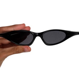 May include: Black sunglasses with a sleek, narrow frame and dark lenses. The sunglasses have a curved design and are held against a white background. The overall style is modern and minimalist.