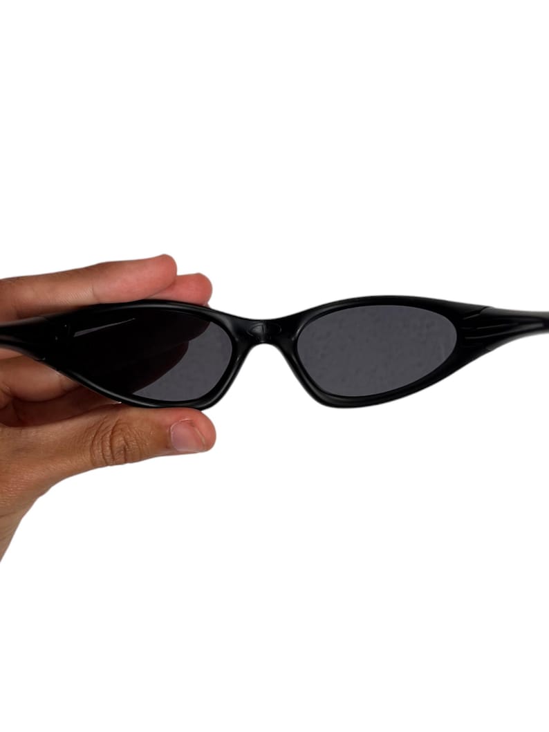May include: Black sunglasses with a sleek, narrow frame and dark lenses. The sunglasses have a curved design and are held against a white background. The overall style is modern and minimalist.