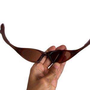 May include: Brown sunglasses with a curved frame and tinted lenses. The sunglasses are held in a hand against a white background. The lenses have a slight oval shape.