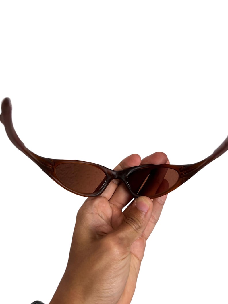 May include: Brown sunglasses with a curved frame and tinted lenses. The sunglasses are held in a hand against a white background. The lenses have a slight oval shape.