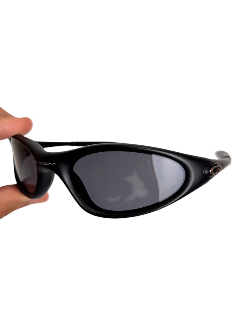 May include: Black sunglasses with a curved, wraparound design. The lenses are dark gray, and the frames are matte black. The Oakley logo is visible on the temple. The sunglasses are held by a hand against a white background.