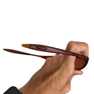 May include: Brown sunglasses with a rectangular frame and tinted lenses. The sunglasses are held up against a white background. The arms of the sunglasses are a darker brown.