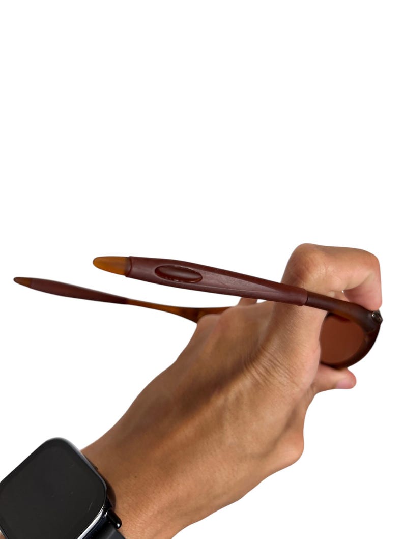 May include: Brown sunglasses with a rectangular frame and tinted lenses. The sunglasses are held up against a white background. The arms of the sunglasses are a darker brown.