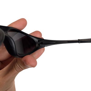 May include: Black sunglasses with dark tinted lenses. The sunglasses have a sleek, modern design with a matte finish. The temples are thin and curve slightly, and the frame is held by a hand.