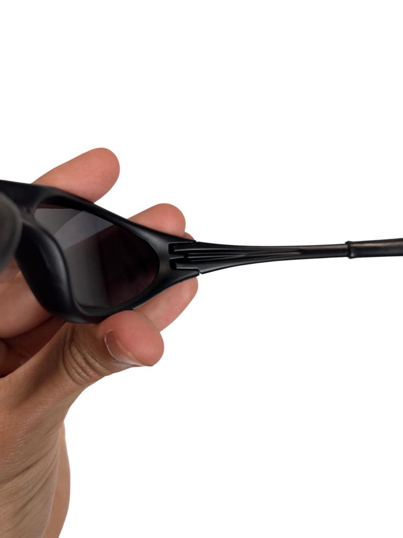 May include: Black sunglasses with dark tinted lenses. The sunglasses have a sleek, modern design with a matte finish. The temples are thin and curve slightly, and the frame is held by a hand.