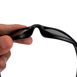 May include: Close-up of black sunglasses with dark lenses. The sunglasses are held up against a white background, showcasing the sleek design and shape of the eyewear. The focus is on the bridge and the top frame of the glasses.