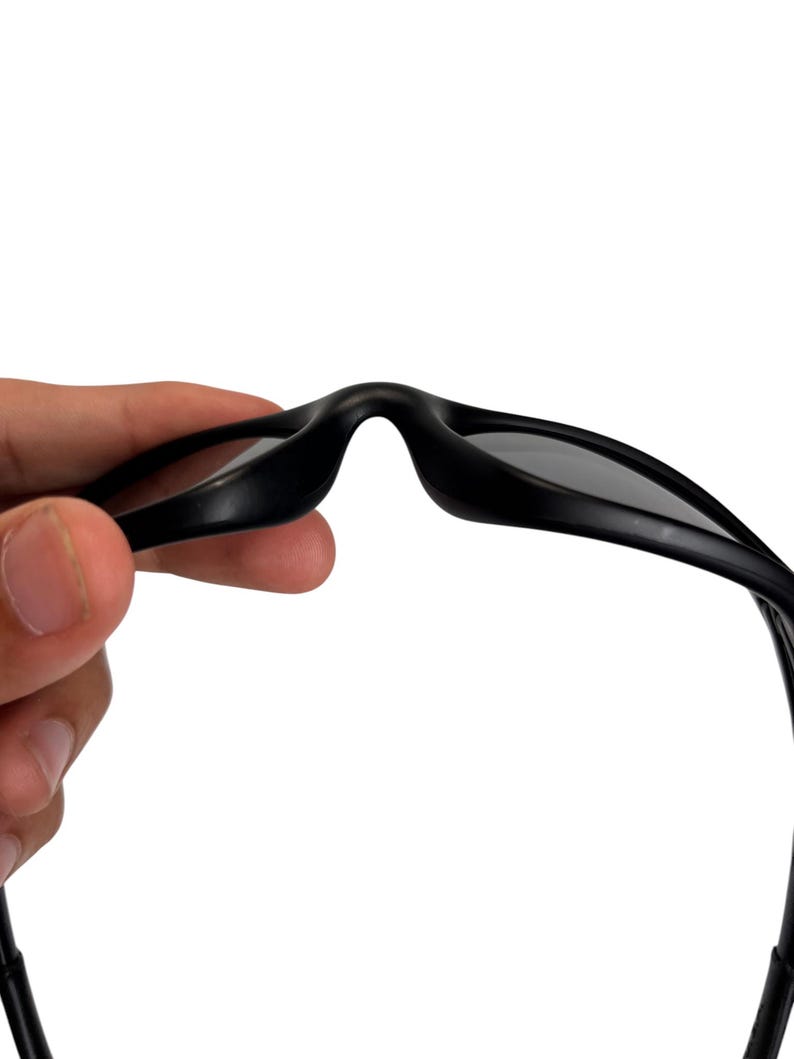 May include: Close-up of black sunglasses with dark lenses. The sunglasses are held up against a white background, showcasing the sleek design and shape of the eyewear. The focus is on the bridge and the top frame of the glasses.