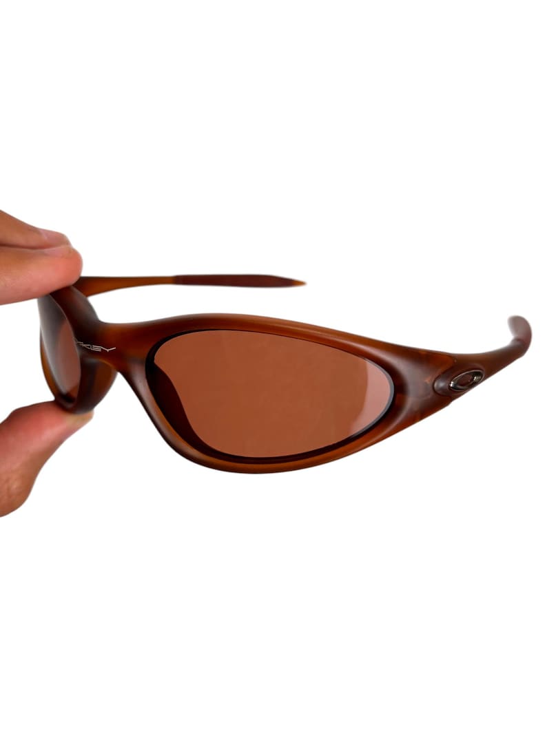 May include: Brown oval-shaped sunglasses with brown lenses. The sunglasses have a curved frame and are held by a hand. The Oakley logo is visible on the frame.