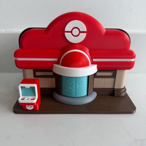 May include: A red and white 3D-printed model of a building with a large Poké Ball symbol on top. The building has a blue door and a small red kiosk. The model sits on a brown base.