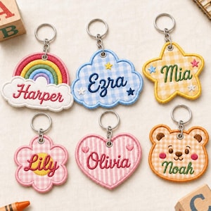 Könnte beinhalten: Sechs bestickte Schlüsselanhänger mit Metallringen in verschiedenen Formen wie Regenbogen, Wolke, Stern, Blume, Herz und Bärengesicht. Jeder Anhänger zeigt einen individuellen Namen: Harper, Ezra, Mia, Lily, Olivia und Noah, gestickt in farbigem Garn.