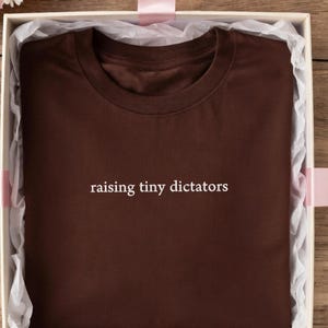 以下が含まれることがあります： 「raising tiny dictators」の白い文字が入った茶色のTシャツ。ピンクのリボンが付いたギフトボックスに入っています。ボックスはピンクと白の花、金のネックレス、木製の表面に散らばる花びらに囲まれています。