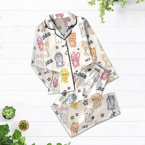 May include: White satin pajama set with a repeating pattern of cartoon animals and stars. The set includes a button-down top with black trim and matching pants. The text "SKZ" is printed throughout the design.
