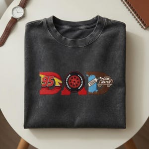 May include: Dark gray t-shirt with the word "DAD" in a Cars-themed design. The "D" features the number 95 in red and yellow, the "A" has a Lightning McQueen tire, and the "D" has Tow Mater.
