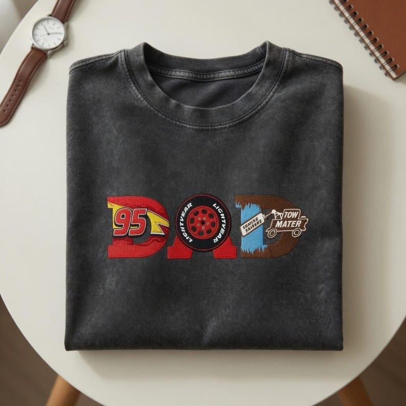 May include: Dark gray t-shirt with the word "DAD" in a Cars-themed design. The "D" features the number 95 in red and yellow, the "A" has a Lightning McQueen tire, and the "D" has Tow Mater.
