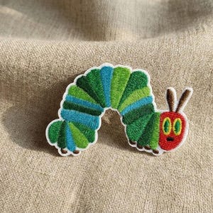 May include: Embroidered patch depicting a colourful caterpillar. The caterpillar has a green and blue segmented body, a red head with brown antennae, and large green eyes. The patch is designed for clothing or accessories.