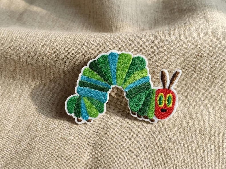 May include: Embroidered patch depicting a colourful caterpillar. The caterpillar has a green and blue segmented body, a red head with brown antennae, and large green eyes. The patch is designed for clothing or accessories.