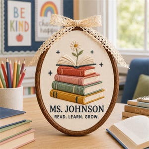 Könnte beinhalten: Ein ovaler Stickrahmen mit einem Stapel aus vier bunten Büchern, einem offenen Buch darauf und einer Gänseblümchenblüte. Der Text lautet MS. JOHNSON und READ. LEARN. GROW. Ein cremefarbenes Spitzenband ist oben am Holzrahmen befestigt.
