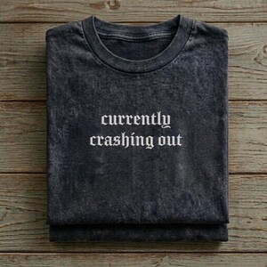 以下が含まれることがあります： 「currently crashing out」の白いゴシック体フォントがプリントされた濃いグレーのTシャツ。Tシャツは折りたたまれており、柔らかく質感のある素材でできており、木製の表面に置かれています。