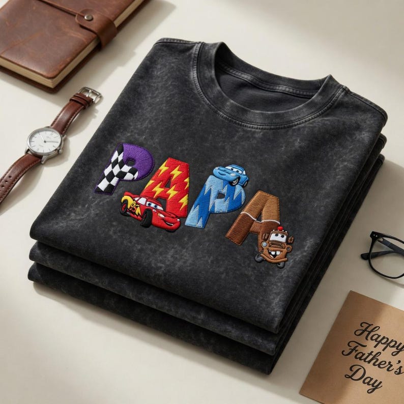 以下が含まれることがあります： カラフルな文字と車のデザインで「PAPA」の刺繍が施された、濃いグレーのTシャツ3枚の重ね着。 ヴィンテージ感のあるウォッシュ加工が施されています。 時計、ノート、そして「Happy Father's Day」と書かれたカードが背景にあります。