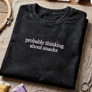 Pode incluir: Camiseta preta com a frase branca "probably thinking about snacks". A camiseta está dobrada sobre uma superfície neutra, com materiais de costura como tesouras e carretéis de linha no fundo.