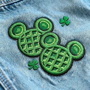 May include: Close-up of a light blue denim jacket with embroidered green Mickey Mouse-shaped waffle patches and two small shamrock patches. The patches have a detailed, textured design with black outlines.