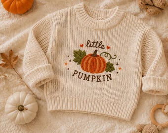 May include: A cream-coloured long-sleeve knit jumper featuring an embroidered orange pumpkin graphic in the centre. The text "little PUMPKIN" is stitched in brown above and below the pumpkin, accented by small red hearts and orange star icons.