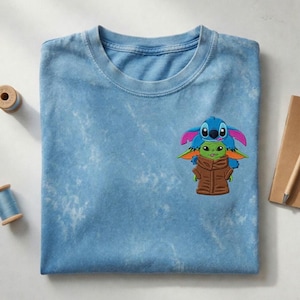 May include: A light blue t-shirt featuring a graphic of Stitch and Grogu. The graphic shows Stitch in blue and pink, and Grogu in green and brown. The shirt is folded on a white surface.