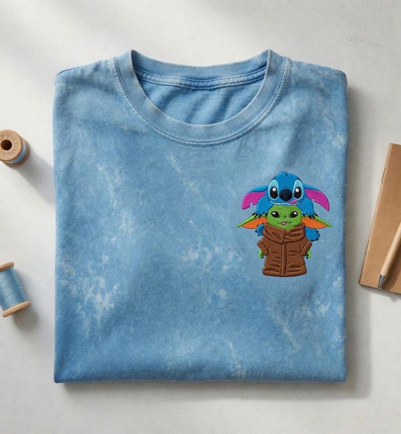 May include: A light blue t-shirt featuring a graphic of Stitch and Grogu. The graphic shows Stitch in blue and pink, and Grogu in green and brown. The shirt is folded on a white surface.