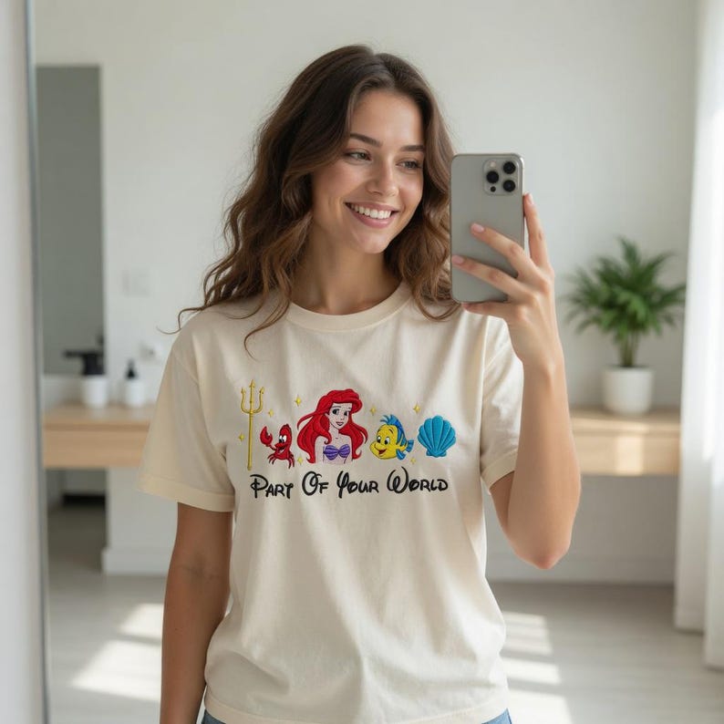 Puede incluir: Camiseta color crema con un dise&ntilde;o bordado colorido de personajes de La Sirenita. El dise&ntilde;o incluye un tridente, un cangrejo, Ariel, Flounder y una concha, con el texto "Part Of Your World" debajo.