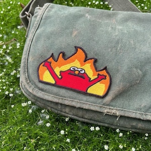 以下が含まれることがあります： フラップとストラップが付いた、使い古された緑色のキャンバスメッセンジャーバッグ。炎に包まれた赤いキャラクターのカラフルな刺繍パッチが付いています。バッグは緑の草と小さな白い花の背景にあります。バッグには使用感があります。
