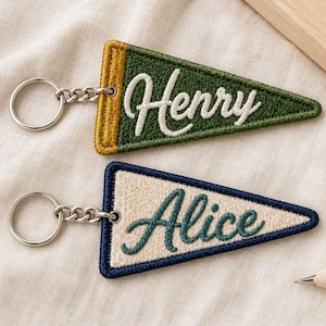 May include: Two triangular embroidered keychains with silver metal rings. The top keychain is dark green with a gold border and the white embroidered name Henry. The bottom keychain is cream with a dark blue border and the dark green embroidered name Alice.
