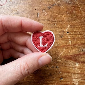 May include: A red heart-shaped embroidered patch with a white border and the letter "L". The patch is held between fingers. Ideal for customising clothing or accessories. The background is a wooden surface.