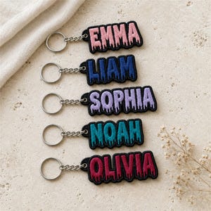 May include: Five embroidered keychains with silver-tone metal rings and chains. Each keychain features a name in a dripping font style with a black border: EMMA in pink, LIAM in blue, SOPHIA in light purple, NOAH in teal, and OLIVIA in dark red.