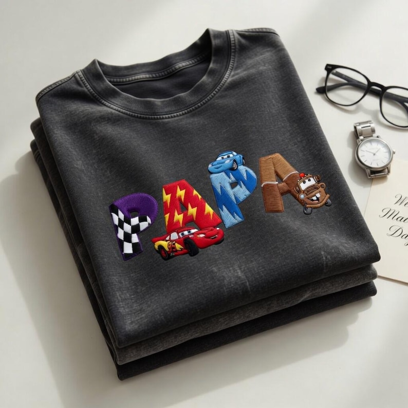 以下が含まれることがあります： 折りたたまれた濃い灰色のTシャツ。漫画風のカラフルな文字で「PAPA」の文字が刺繍されています。文字にはレーシングカーとチェッカーフラッグの画像が使用されています。シャツはきれいに折りたたまれており、新品であることがわかります。