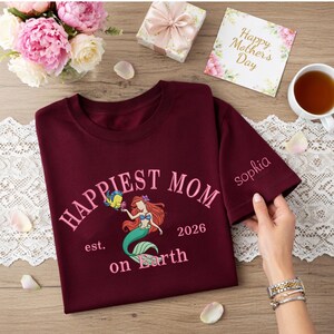 May include: Burgundy t-shirt with the words "Happiest Mom on Earth" and a mermaid graphic. The sleeve is embroidered with the name "Sophia". A "Happy Mother's Day" card and flowers are in the background.