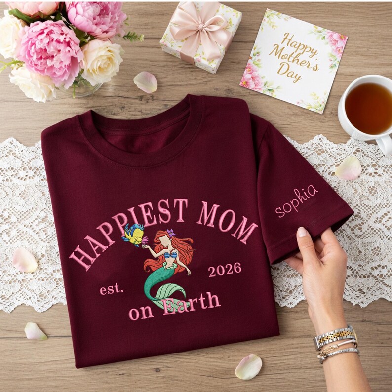 May include: Burgundy t-shirt with the words "Happiest Mom on Earth" and a mermaid graphic. The sleeve is embroidered with the name "Sophia". A "Happy Mother's Day" card and flowers are in the background.