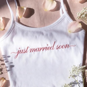 May include: A white crop top with thin straps, featuring the embroidered phrase "just married soon" in a cursive script. The top is adorned with pink rose petals and small white flowers, set on a pink backdrop.