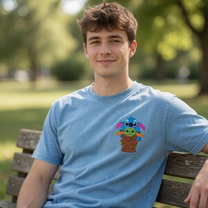 May include: Pale blue t-shirt with a colourful graphic of Stitch and Grogu. The design shows Stitch on top of Grogu, both wearing brown jackets. The shirt has a crew neck and short sleeves.