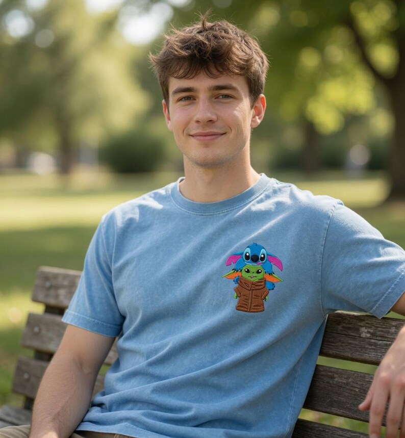 May include: Pale blue t-shirt with a colourful graphic of Stitch and Grogu. The design shows Stitch on top of Grogu, both wearing brown jackets. The shirt has a crew neck and short sleeves.