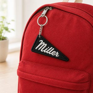 May include: A deep red canvas backpack featuring a silver metal zipper and a black triangular pennant keychain attached to the zipper pull. The pennant is embroidered with the white cursive text Miller and has a black stitched border.