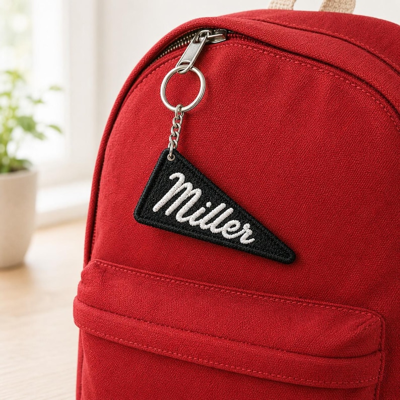 May include: A deep red canvas backpack featuring a silver metal zipper and a black triangular pennant keychain attached to the zipper pull. The pennant is embroidered with the white cursive text Miller and has a black stitched border.