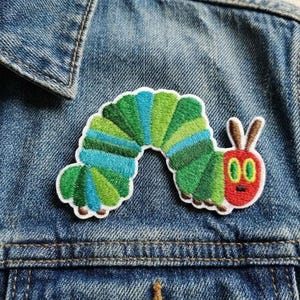 May include: Embroidered patch featuring a colorful caterpillar design with green, blue, and red accents. The patch is attached to a denim jacket, showcasing its vibrant colors and detailed stitching. A fun, whimsical accessory.