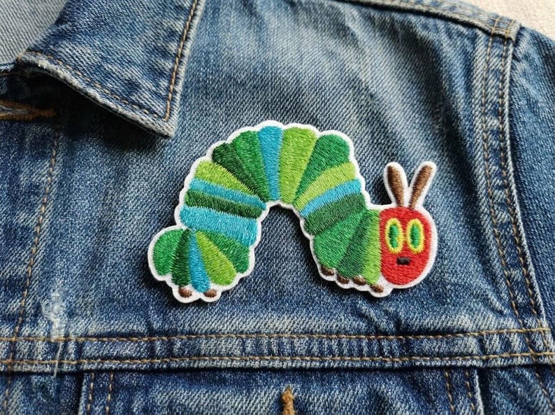 May include: Embroidered patch of a caterpillar in green, blue, and red, attached to a denim jacket. The patch's design is detailed, with a white border. A colourful, whimsical accessory for clothing or bags.