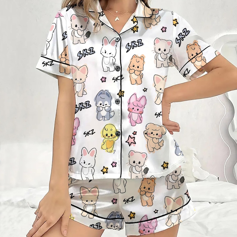 May include: White satin pajama set with a repeating pattern of cartoon animals and stars. The short-sleeved top and shorts are trimmed in black and feature the text "SKZ". The set includes a button-down shirt and shorts.