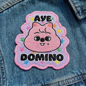 Pu&ograve; includere: Toppa ricamata con un personaggio dei cartoni animati rosa e la scritta "AYE DOMINO" in nero. La toppa ha un bordo lilla chiaro con accenti di stelle colorate ed &egrave; cucita su una giacca di jeans.