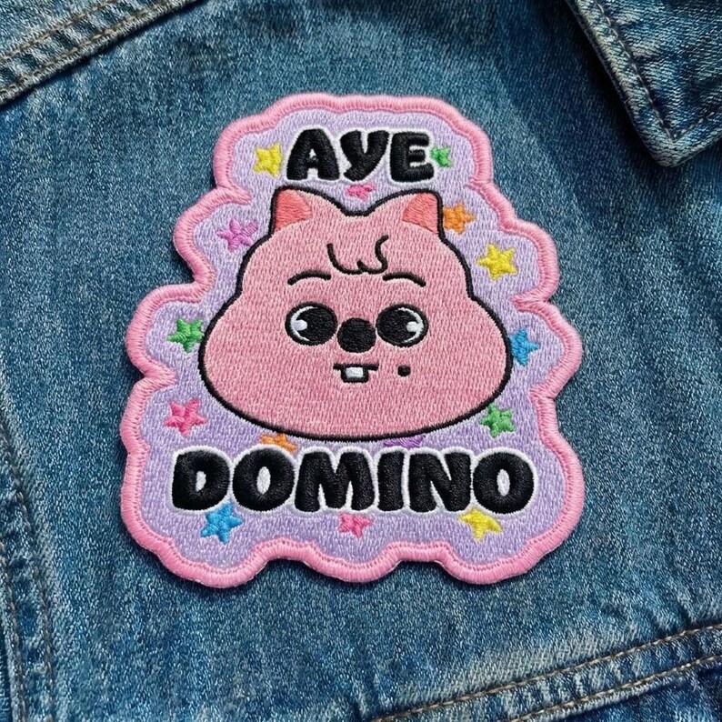 Pu&ograve; includere: Toppa ricamata con un personaggio dei cartoni animati rosa e la scritta "AYE DOMINO" in nero. La toppa ha un bordo lilla chiaro con accenti di stelle colorate ed &egrave; cucita su una giacca di jeans.