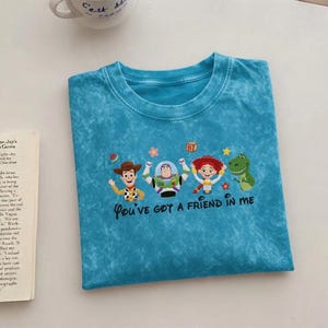 Puede incluir: Camiseta turquesa con un dise&ntilde;o de personajes de Toy Story: Woody, Buzz Lightyear, Jessie y Rex. El texto "You've Got a Friend in Me" est&aacute; debajo. La camiseta tiene cuello redondo y un aspecto descolorido.