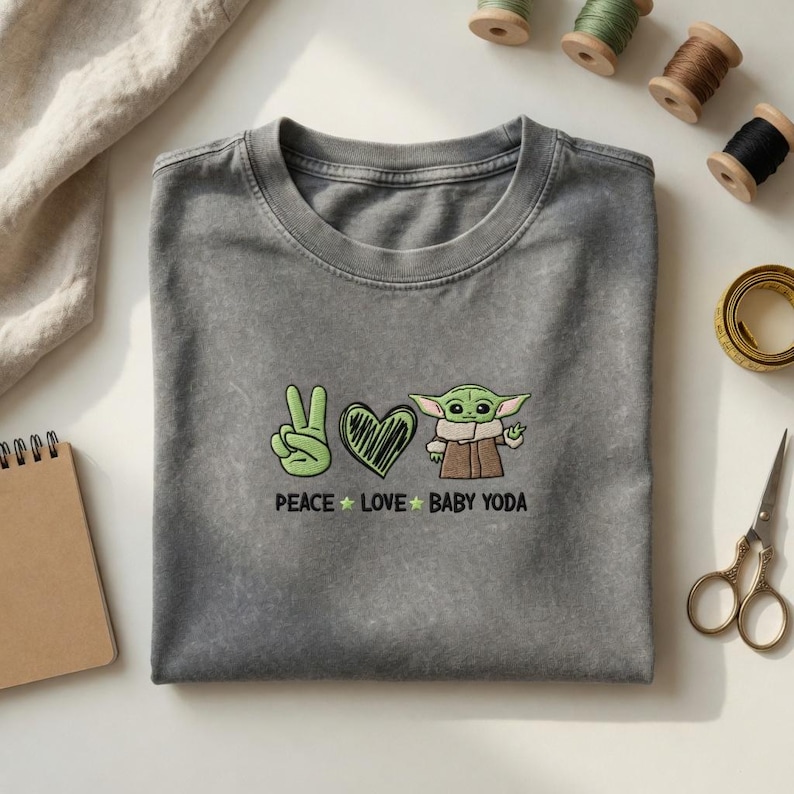 Może przedstawiać: Szara koszulka z haftowanym wzorem przedstawiającym znak pokoju, serce i Baby Yodę. Tekst "PEACE LOVE BABY YODA" znajduje się poniżej. Widoczne są akcesoria do szycia, w tym szpulki nici i nożyczki.